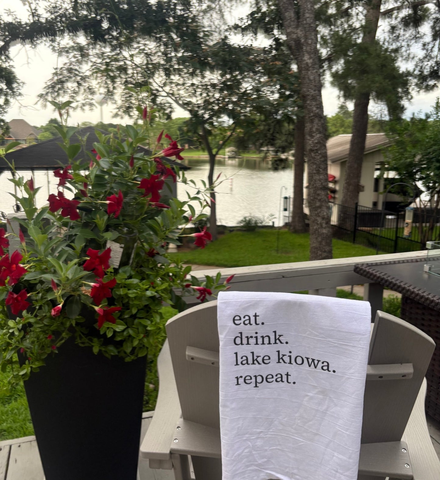 Outdoor setting with a sign reading 'eat. drink. lake kiowa. repeat.' near flowers and a lake.