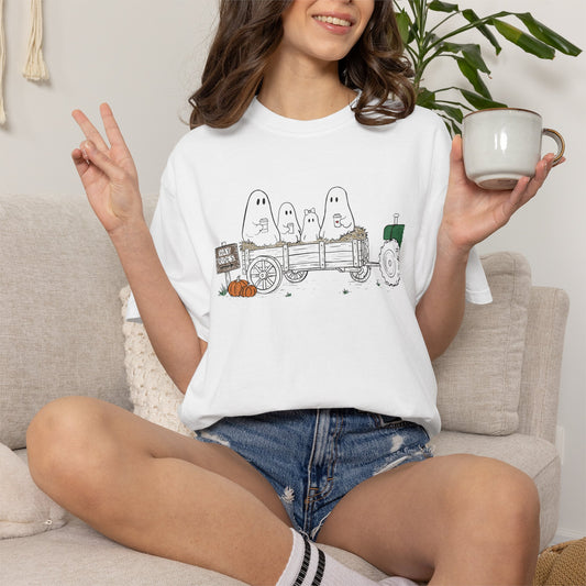 Comfort Colors white t-shirt featuring a hand-drawn ghost family riding in a hay wagon with pumpkins, coffee mugs, and a vintage tractor from The Story of the Ghost Chapter 1 collection.