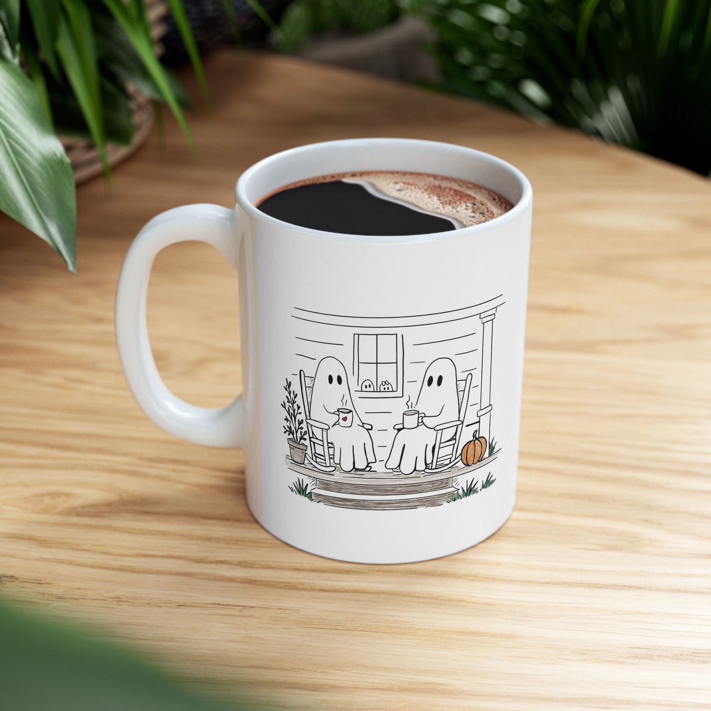 White ceramic mug featuring a hand-drawn ghost family sitting on a front porch in rocking chairs with coffee, with two little ghosts peeking through the window and subtle fall color accents.