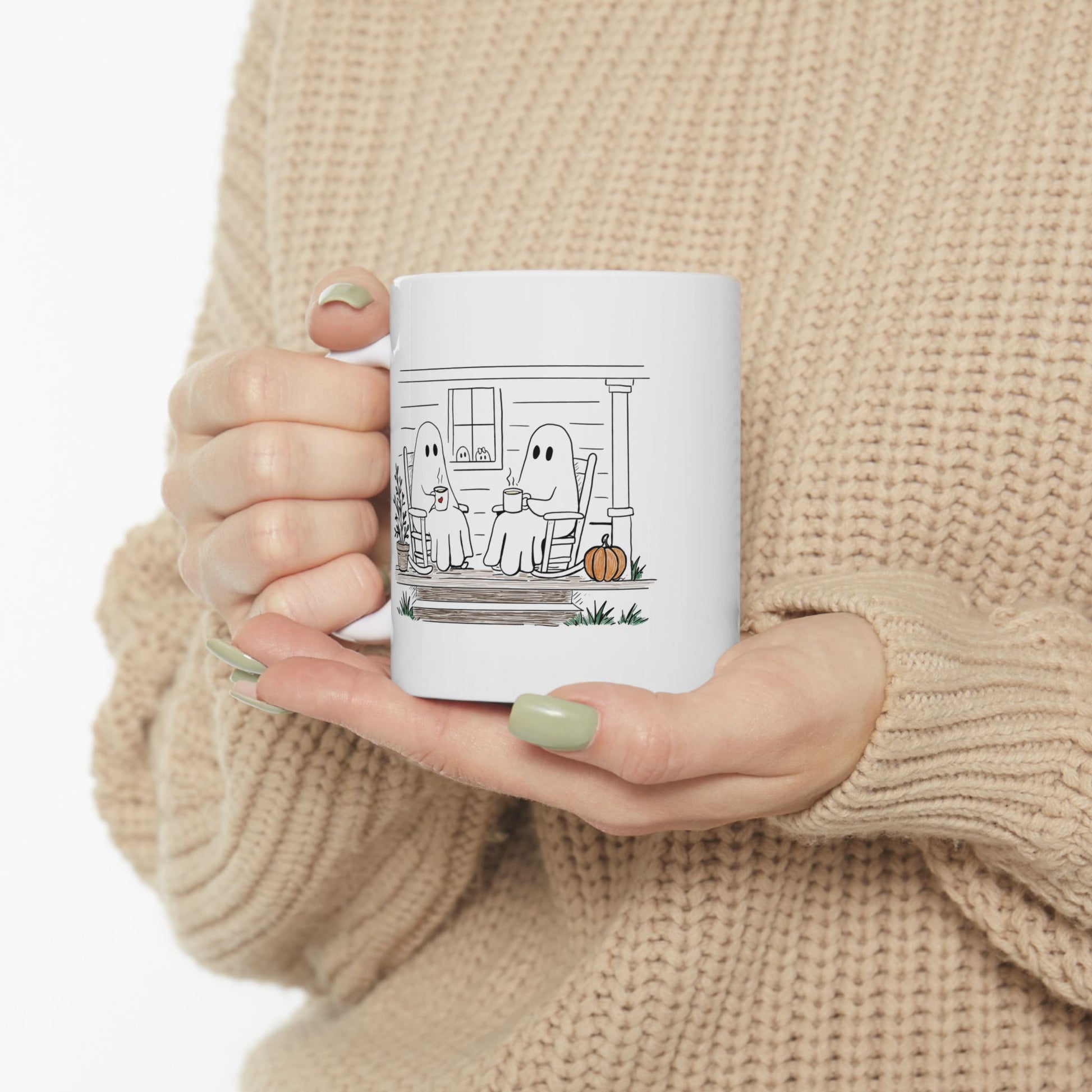 White ceramic mug featuring a hand-drawn ghost family sitting on a front porch in rocking chairs with coffee, with two little ghosts peeking through the window and subtle fall color accents.
