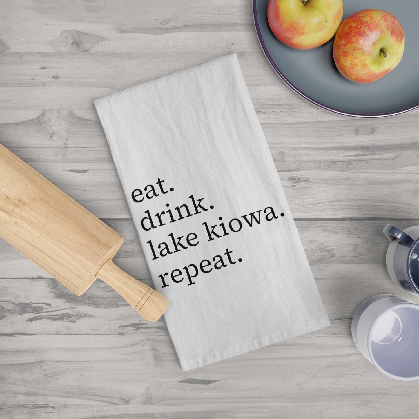 Kitchen towel with 'eat. drink. lake kiowa. repeat.' text on a wooden surface with apples and a rolling pin.