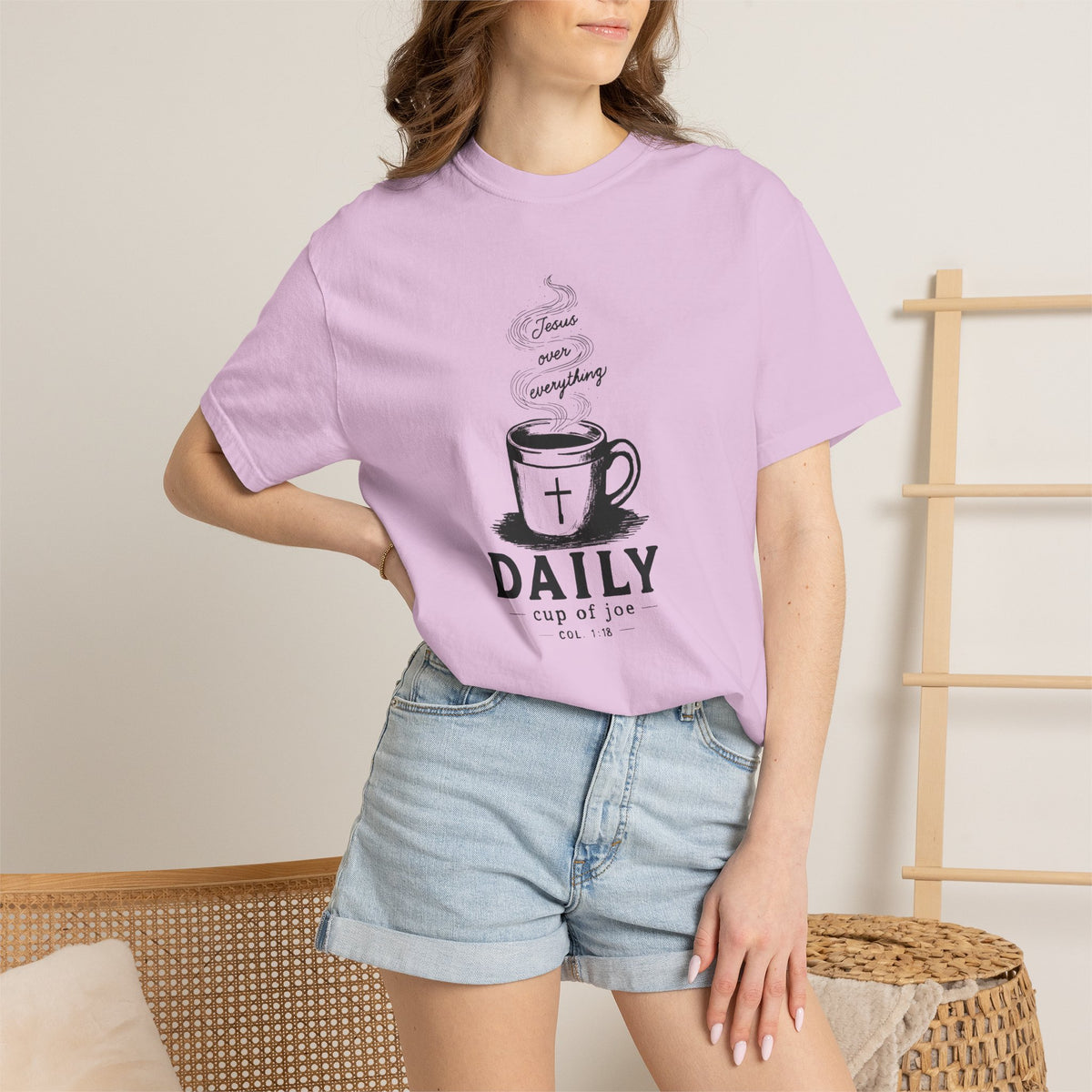 A woman wearing a light purple graphic t-shirt with a black coffee cup design and the text 'DAILY cup of joe' and 'Jesus Over Everything', paired with light blue denim shorts in a warm indoor setting.