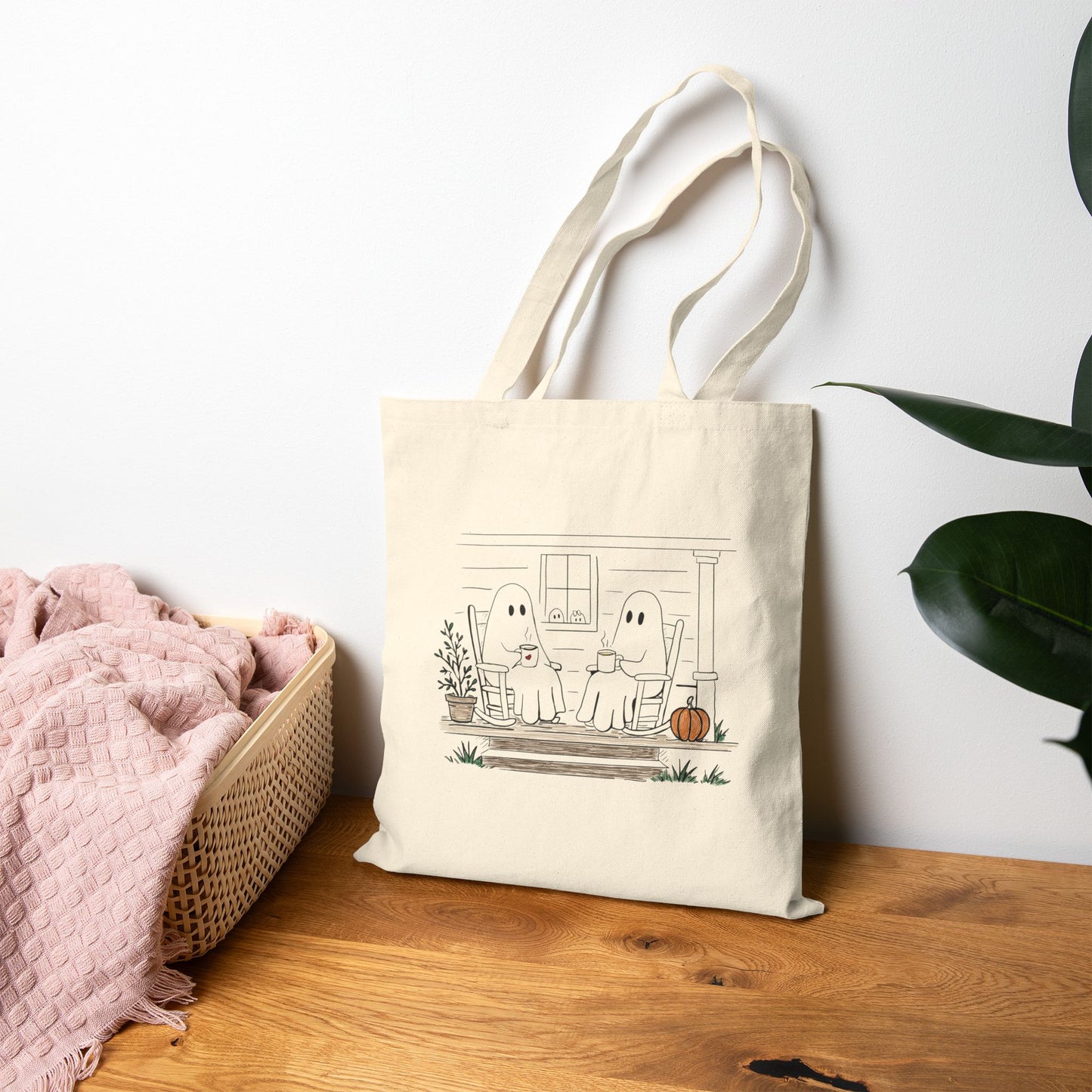 Natural canvas tote bag featuring a hand-drawn ghost family relaxing with coffee in rocking chairs on a front porch, with two little ghosts peeking through the window, a pumpkin, and subtle fall accents.
