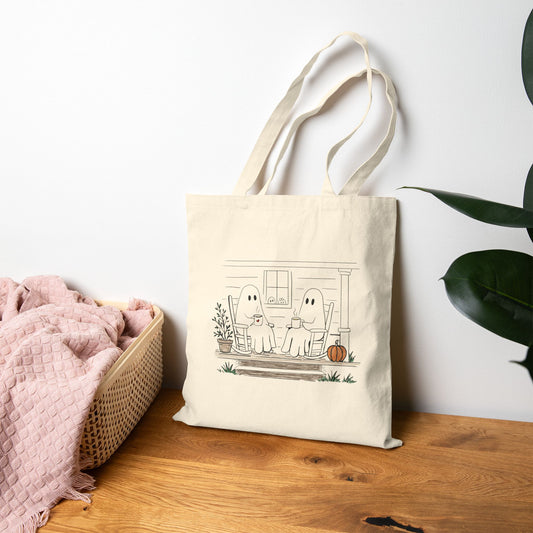 Natural canvas tote bag featuring a hand-drawn ghost family relaxing with coffee in rocking chairs on a front porch, with two little ghosts peeking through the window, a pumpkin, and subtle fall accents.