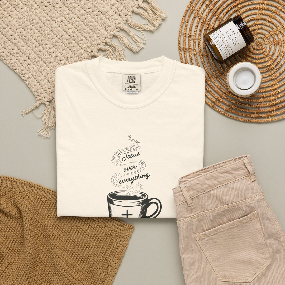 A folded white crewneck t-shirt with a “Jesus Over Everything” graphic on the front, styled in a flat lay with beige pants, a woven blanket, a candle jar, and a small cup on a neutral background.