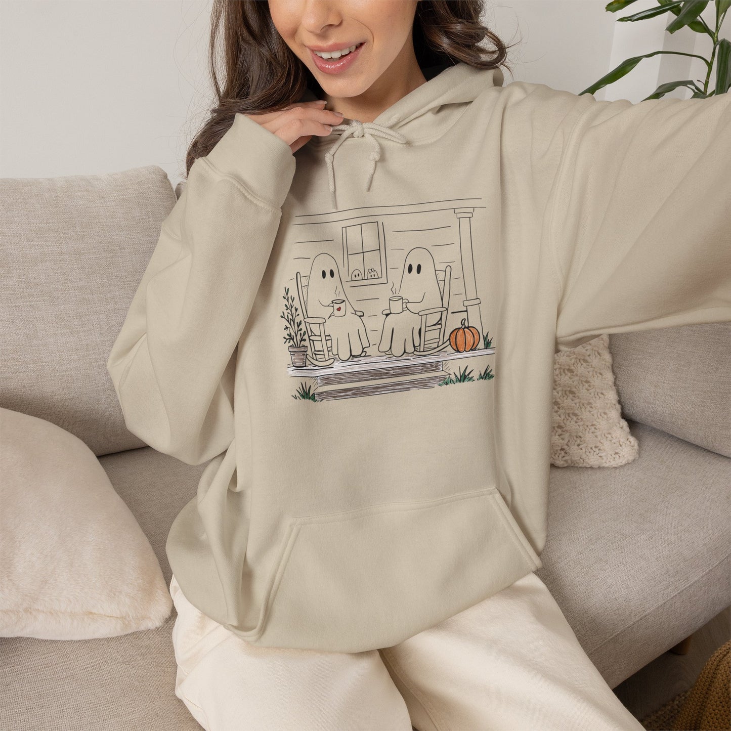 Pullover hoodie featuring a hand-drawn ghost family enjoying coffee in rocking chairs on a front porch, with two little ghosts peeking through the window, a pumpkin, and subtle fall color accents.