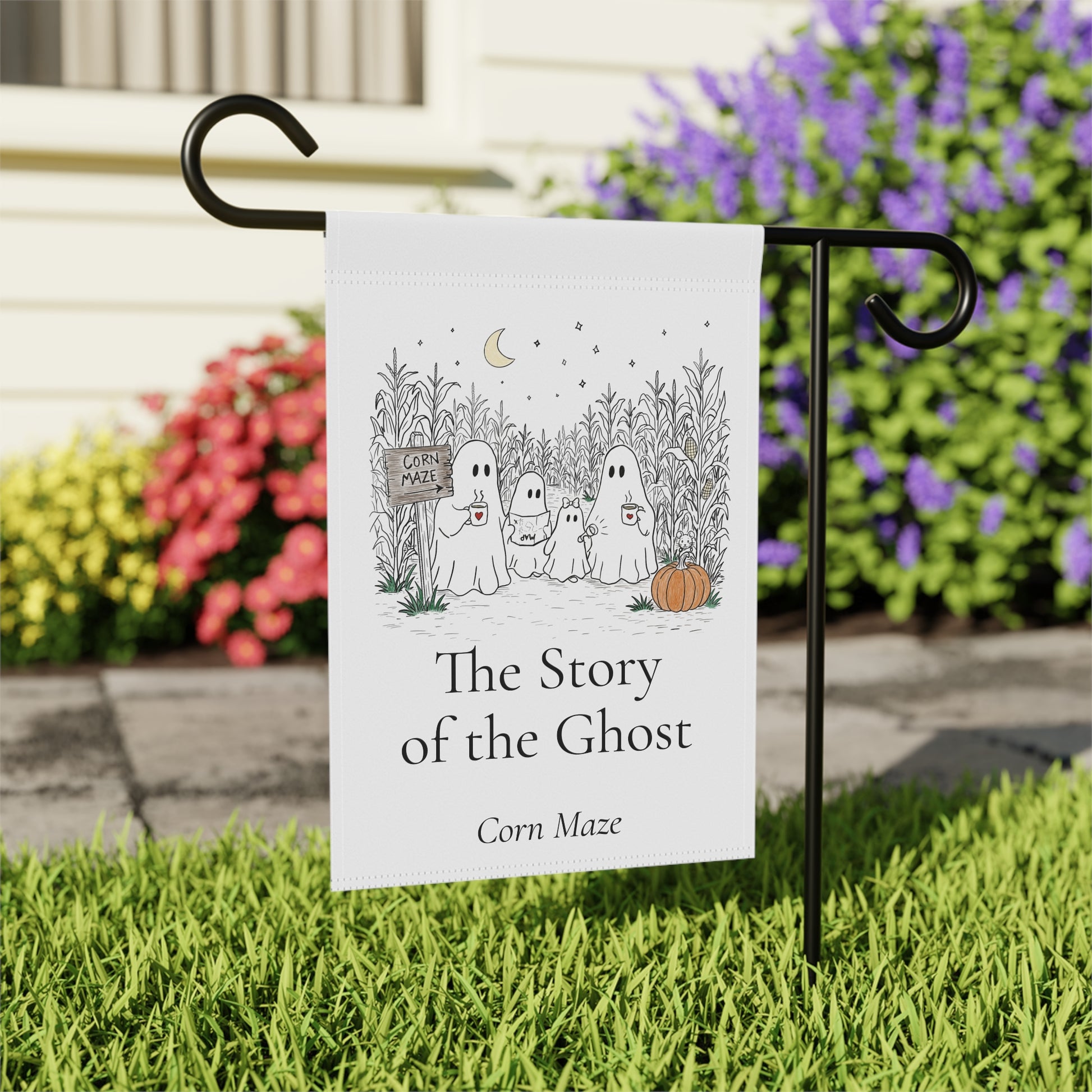 A white garden flag with a black metal stand, featuring a ghostly family illustration and the text 'The Story of the Ghost' and 'Corn Maze'. The flag is displayed on a black H-shaped stand in a grassy yard with colorful flowers in the background.
