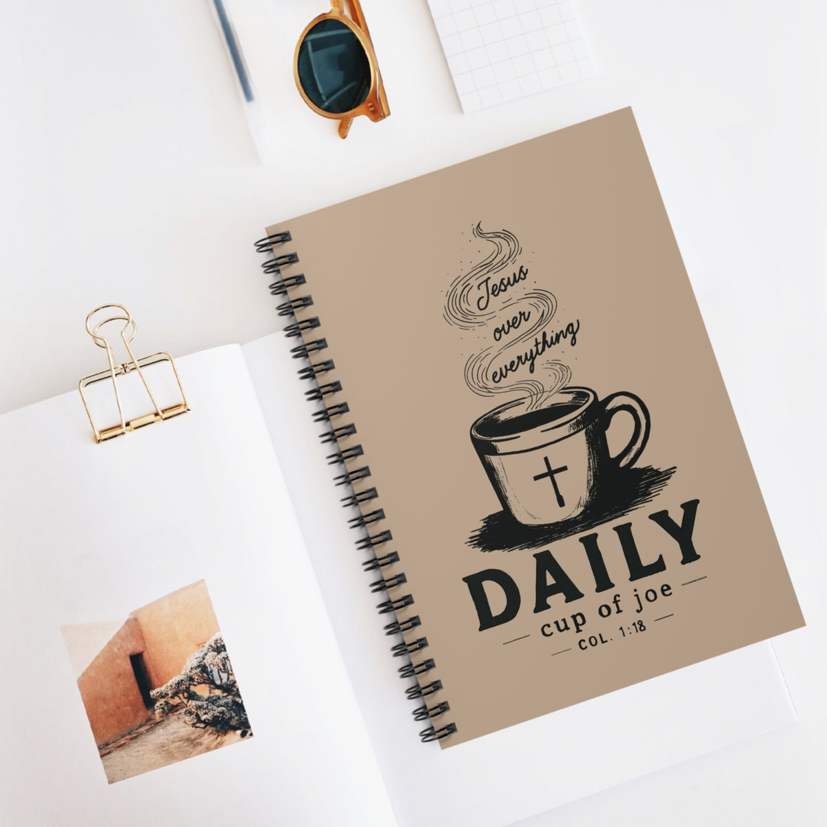 A spiral-bound daily journal with a beige cover lies open on a white desk, surrounded by styled props including sunglasses, a gold binder clip, and a small photo print. The cover features a black coffee cup illustration with a cross and the text 'Jesus over everything' and 'DAILY cup of joe'.