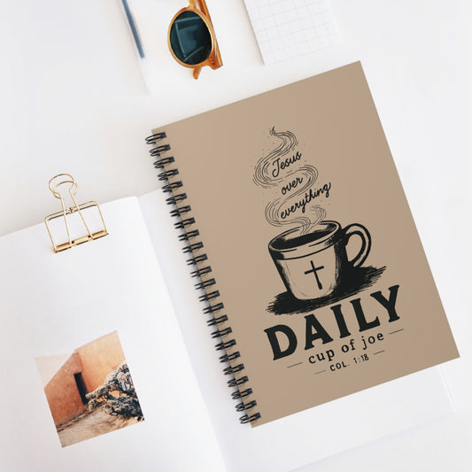 A spiral-bound daily journal with a beige cover lies open on a white desk, surrounded by styled props including sunglasses, a gold binder clip, and a small photo print. The cover features a black coffee cup illustration with a cross and the text 'Jesus over everything' and 'DAILY cup of joe'.