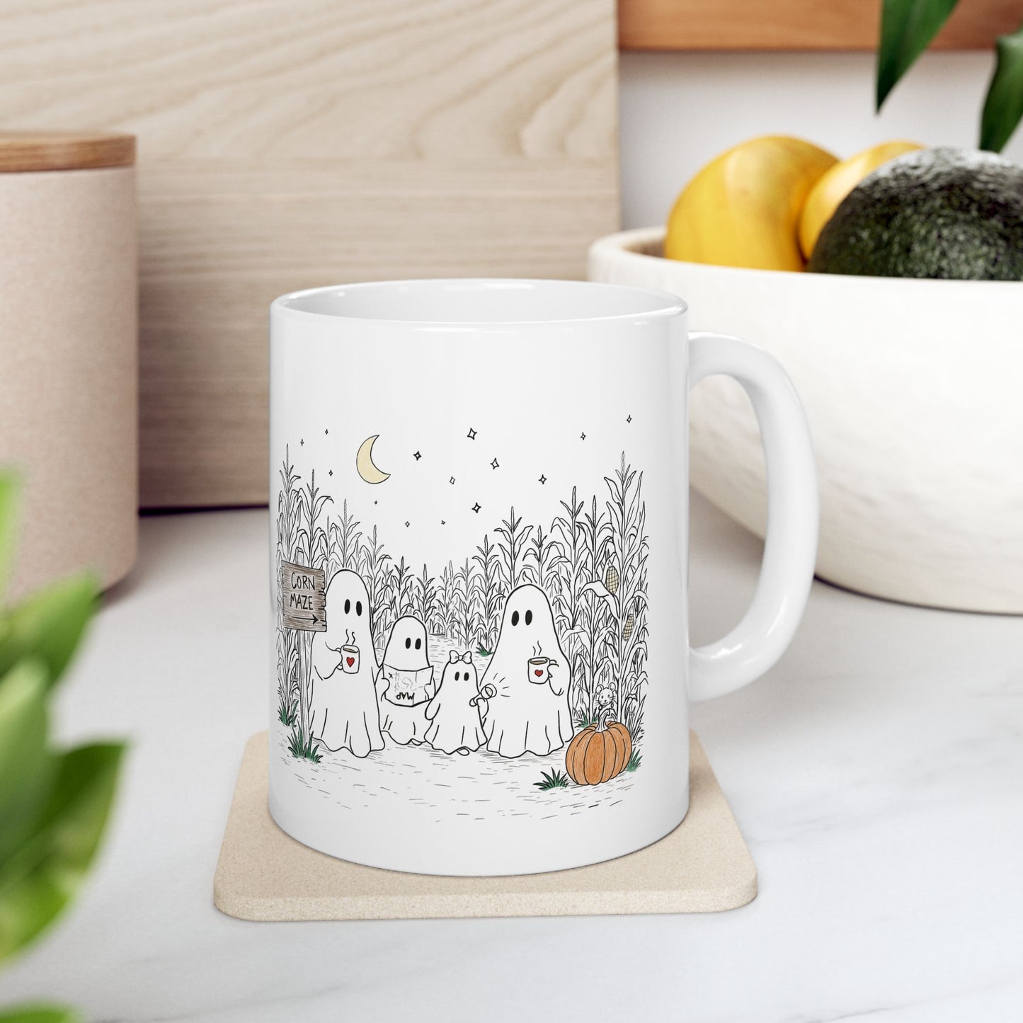 A white ceramic mug with a cute cartoon ghost family illustration in a cornfield, placed on a light wood coaster in a kitchen setting.