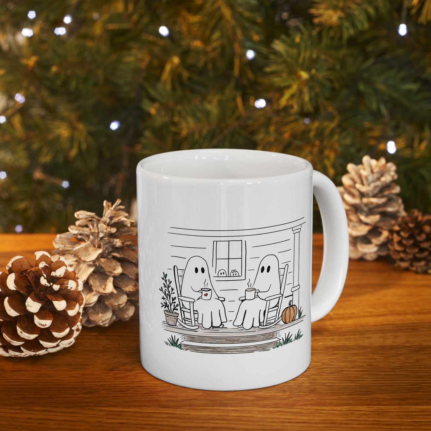 White ceramic mug featuring a hand-drawn ghost family sitting on a front porch in rocking chairs with coffee, with two little ghosts peeking through the window and subtle fall color accents.