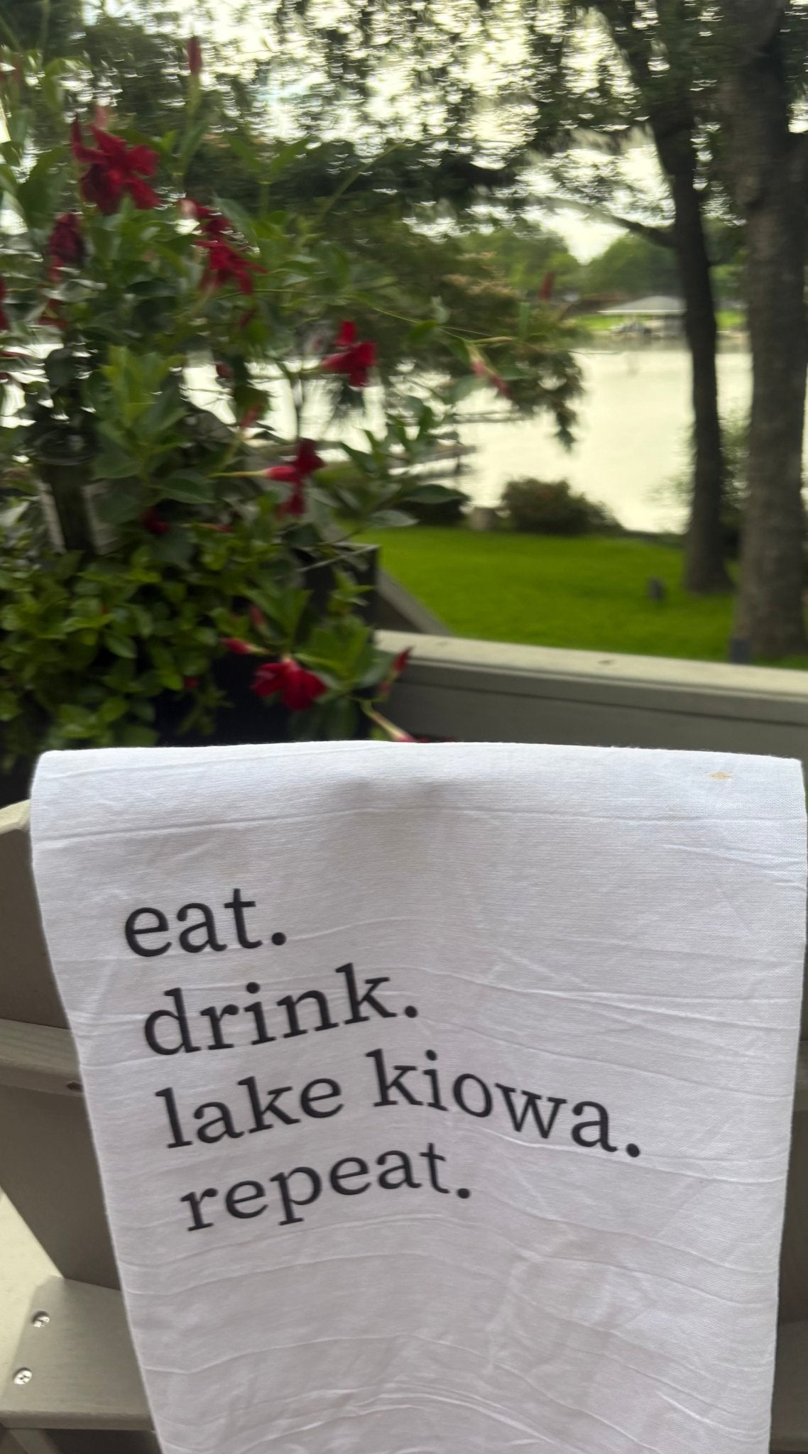 Tea towel with 'eat. drink. lake kiowa. repeat.' text on a window sill with outdoor view.
