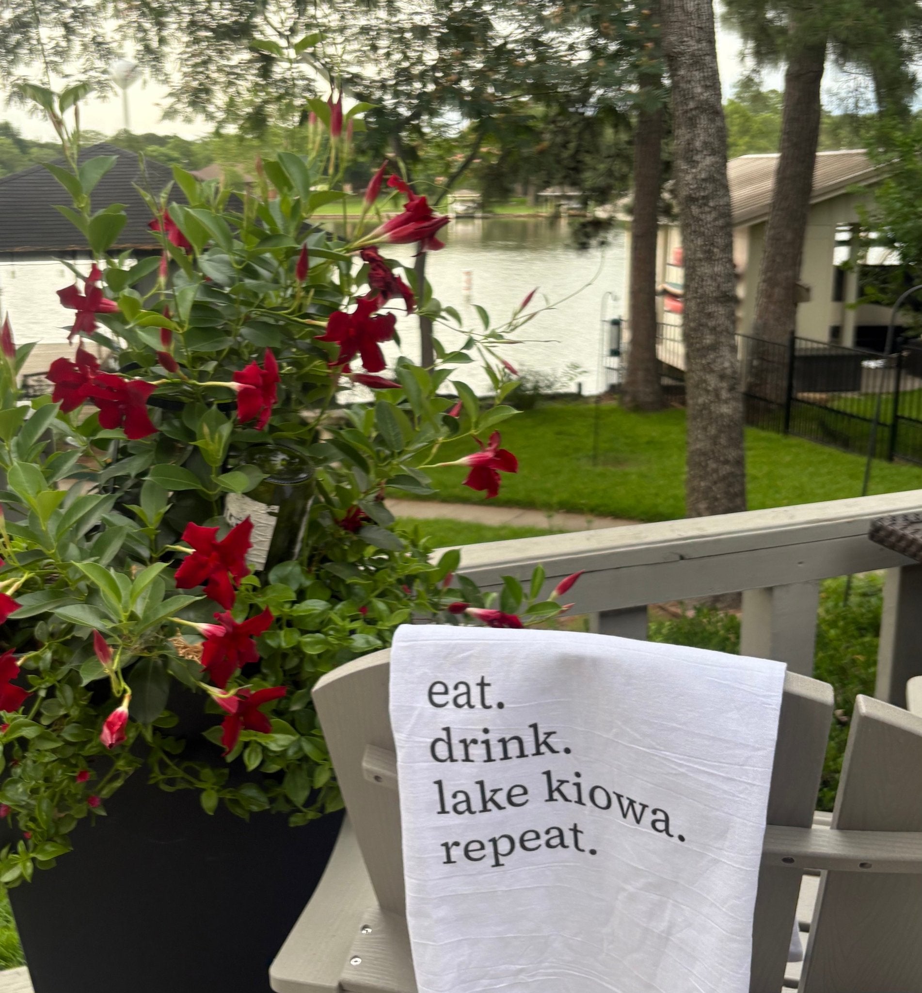 Chair with a towel reading 'eat. drink. lake kiowa. repeat.' next to potted red flowers on a deck.
