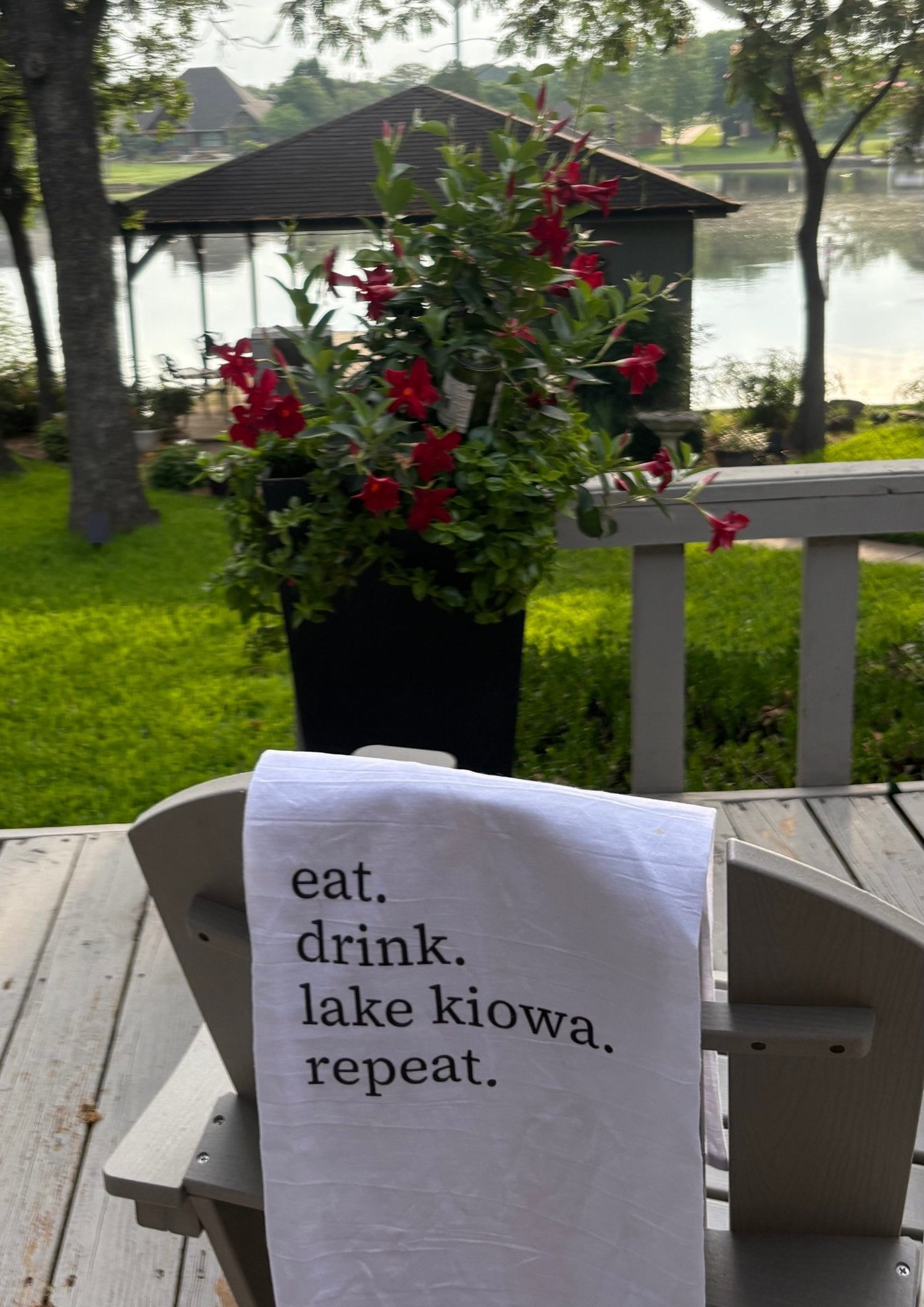 Towel with 'eat. drink. lake kiowa. repeat.' text on a wooden deck with flowers and gazebo in the background.