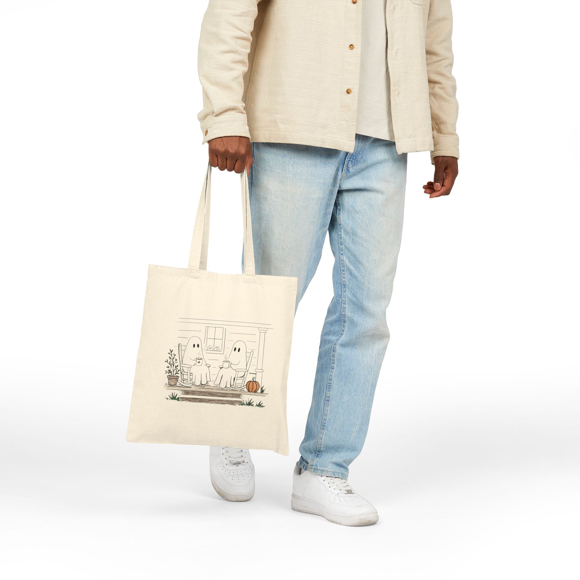 Natural canvas tote bag featuring a hand-drawn ghost family relaxing with coffee in rocking chairs on a front porch, with two little ghosts peeking through the window, a pumpkin, and subtle fall accents.