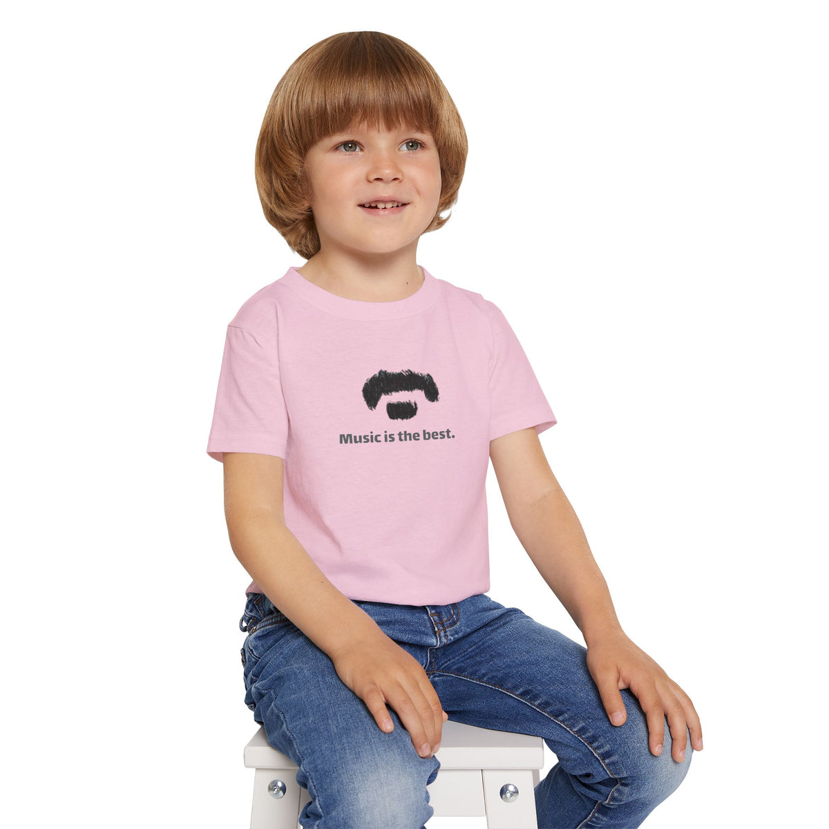 A young boy with light brown hair sits on a small white stool wearing a light pink t-shirt with a black graphic of a mustached face and the text 'Music is the best.' He is also wearing blue jeans against a solid white background.