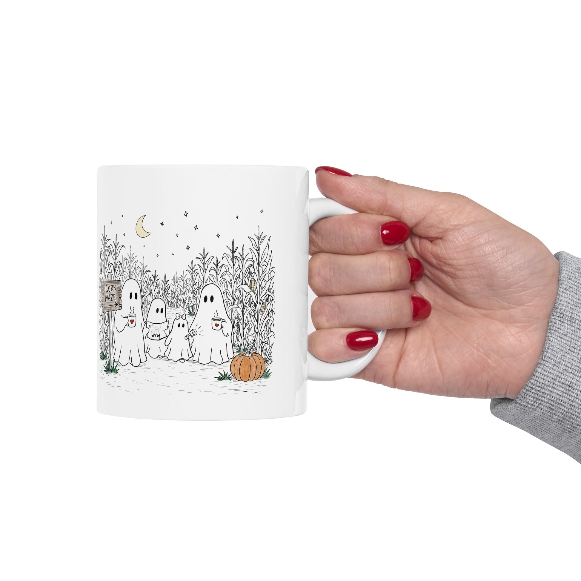 A white ceramic mug with a cute cartoon illustration of four ghosts in a cornfield at night, held by a hand with red-painted nails against a plain white background.