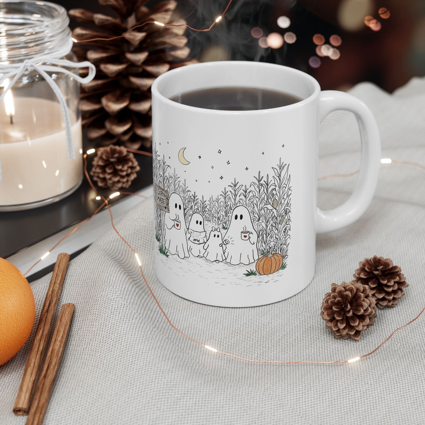 A white ceramic coffee mug with a cute cartoon ghost family illustration, filled with black coffee and set on a light fabric surface surrounded by autumn-themed props like pinecones, a candle, cinnamon sticks, and fairy lights.