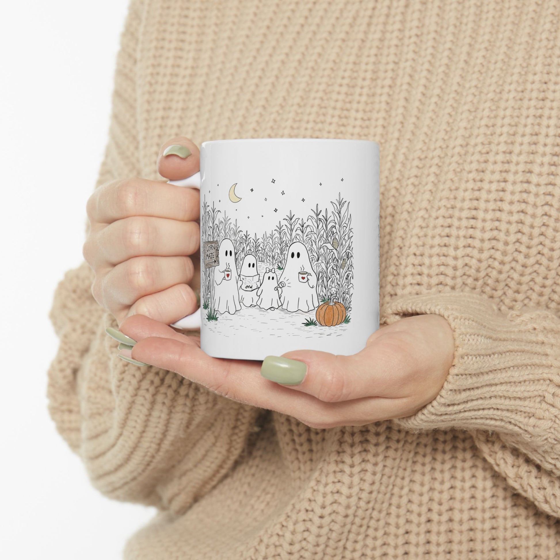A person in a beige knit sweater holds a white ceramic mug with a cute ghost illustration on a white background.