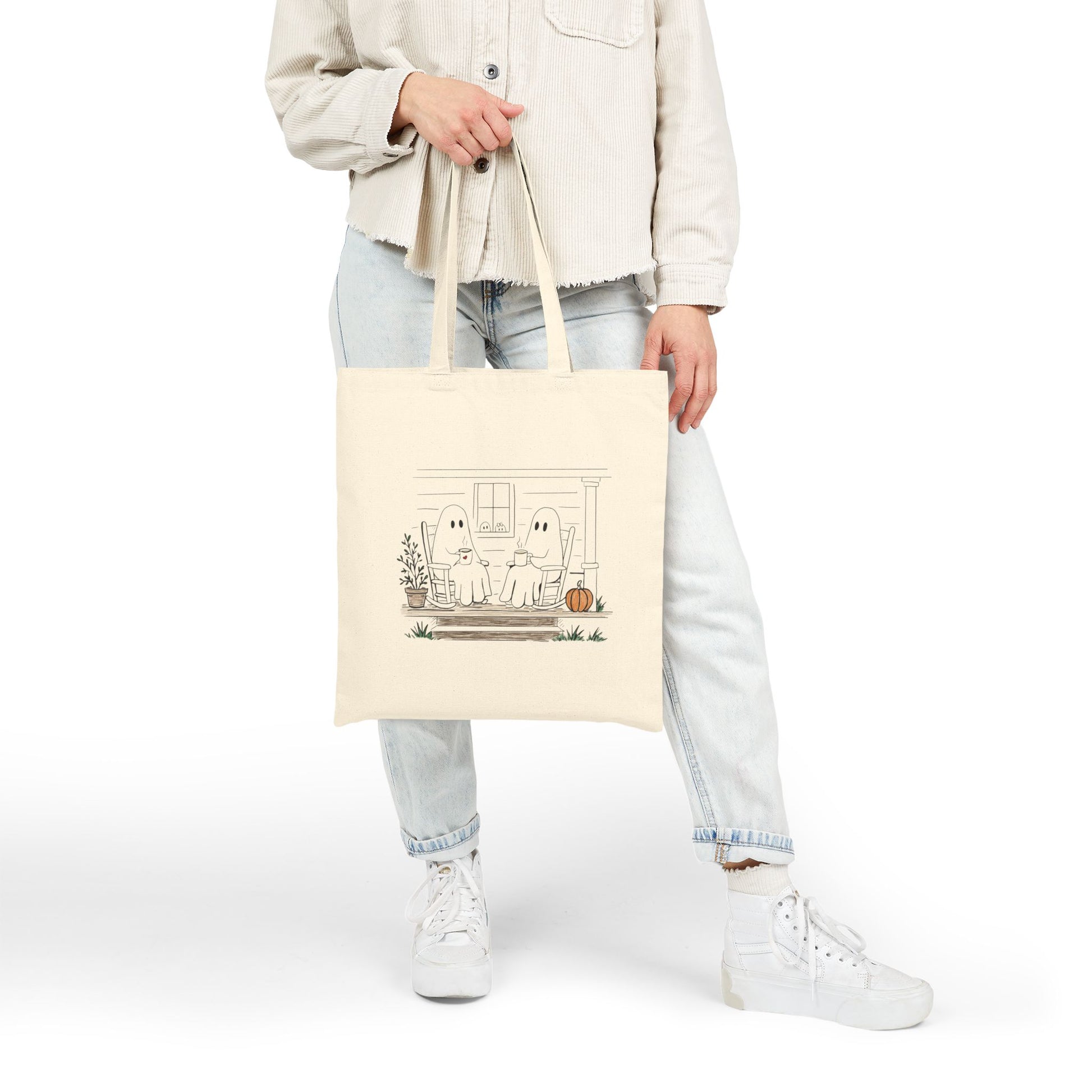 Natural canvas tote bag featuring a hand-drawn ghost family relaxing with coffee in rocking chairs on a front porch, with two little ghosts peeking through the window, a pumpkin, and subtle fall accents.