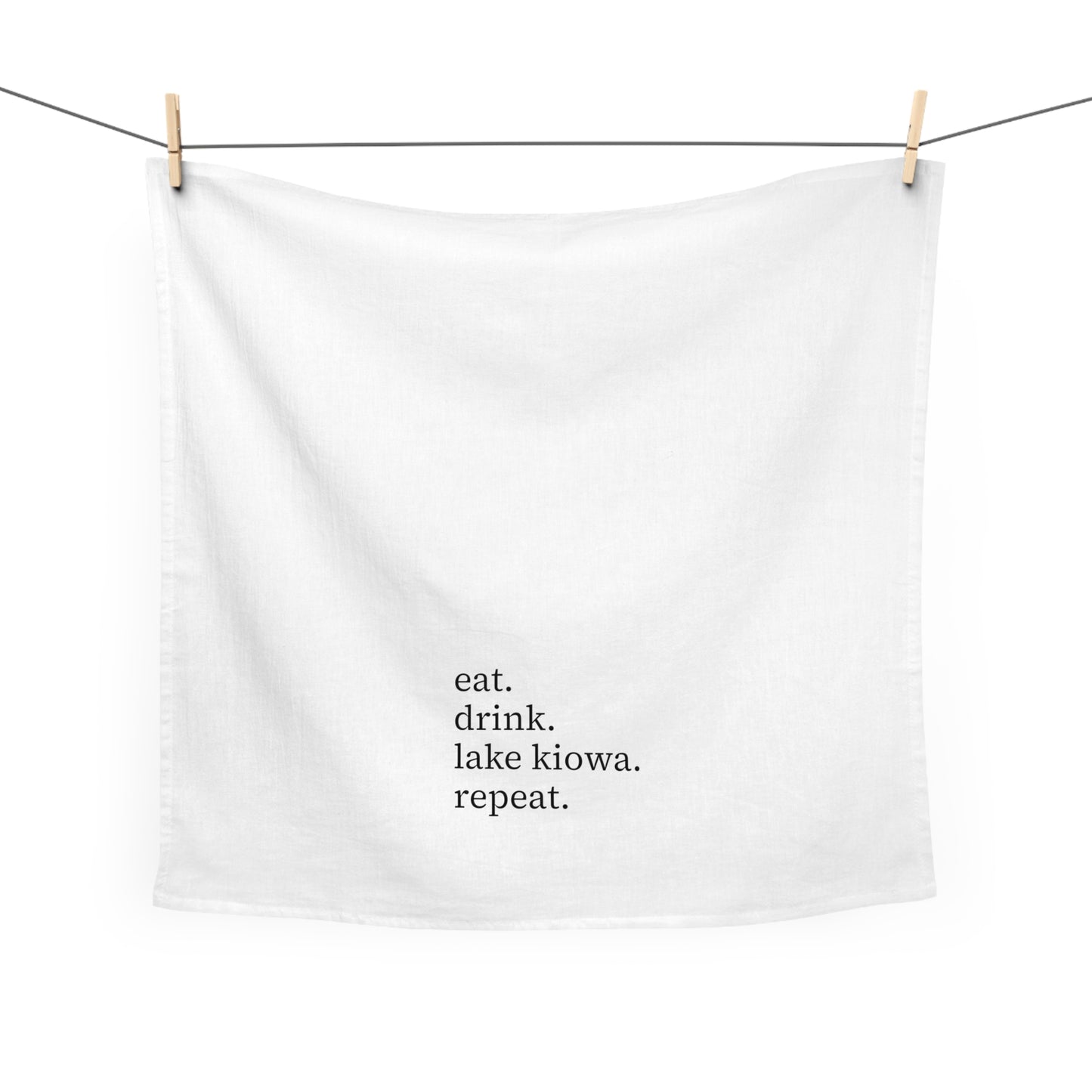White towel with text 'eat. drink. lake kiowa. repeat.' hanging on a clothesline.
