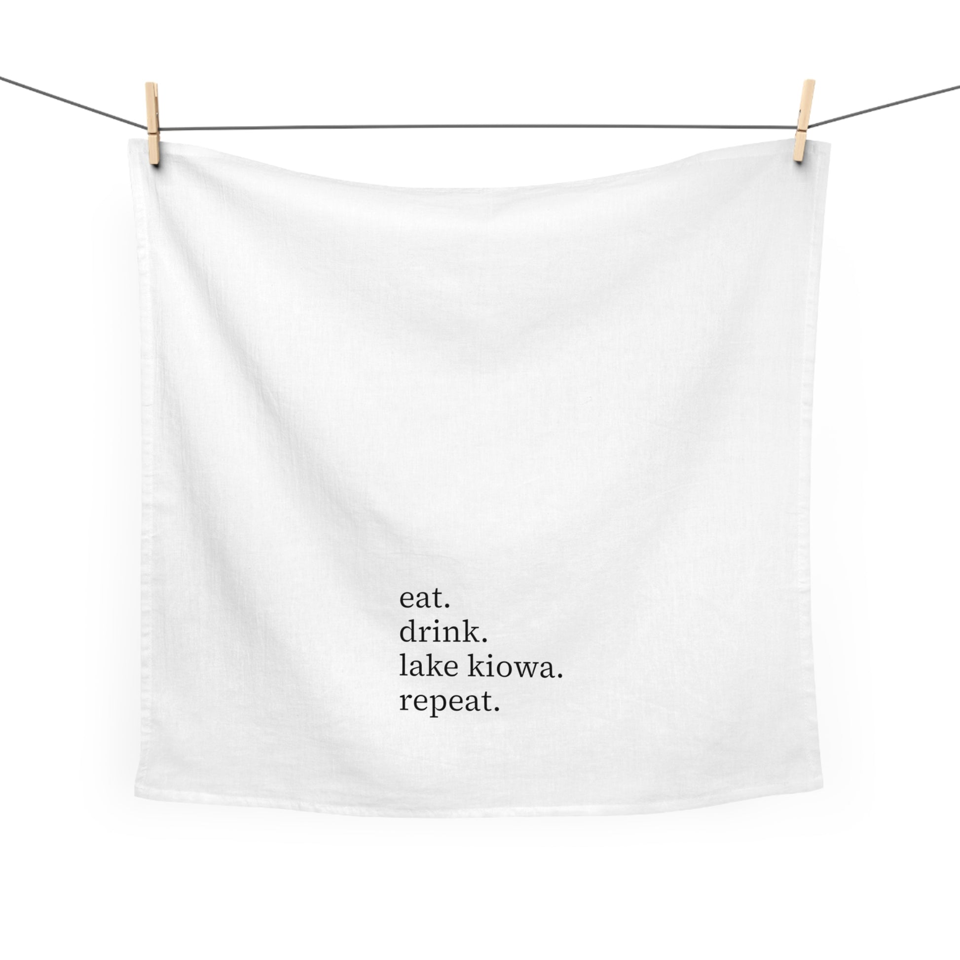 White towel with text 'eat. drink. lake kiowa. repeat.' hanging on a clothesline.