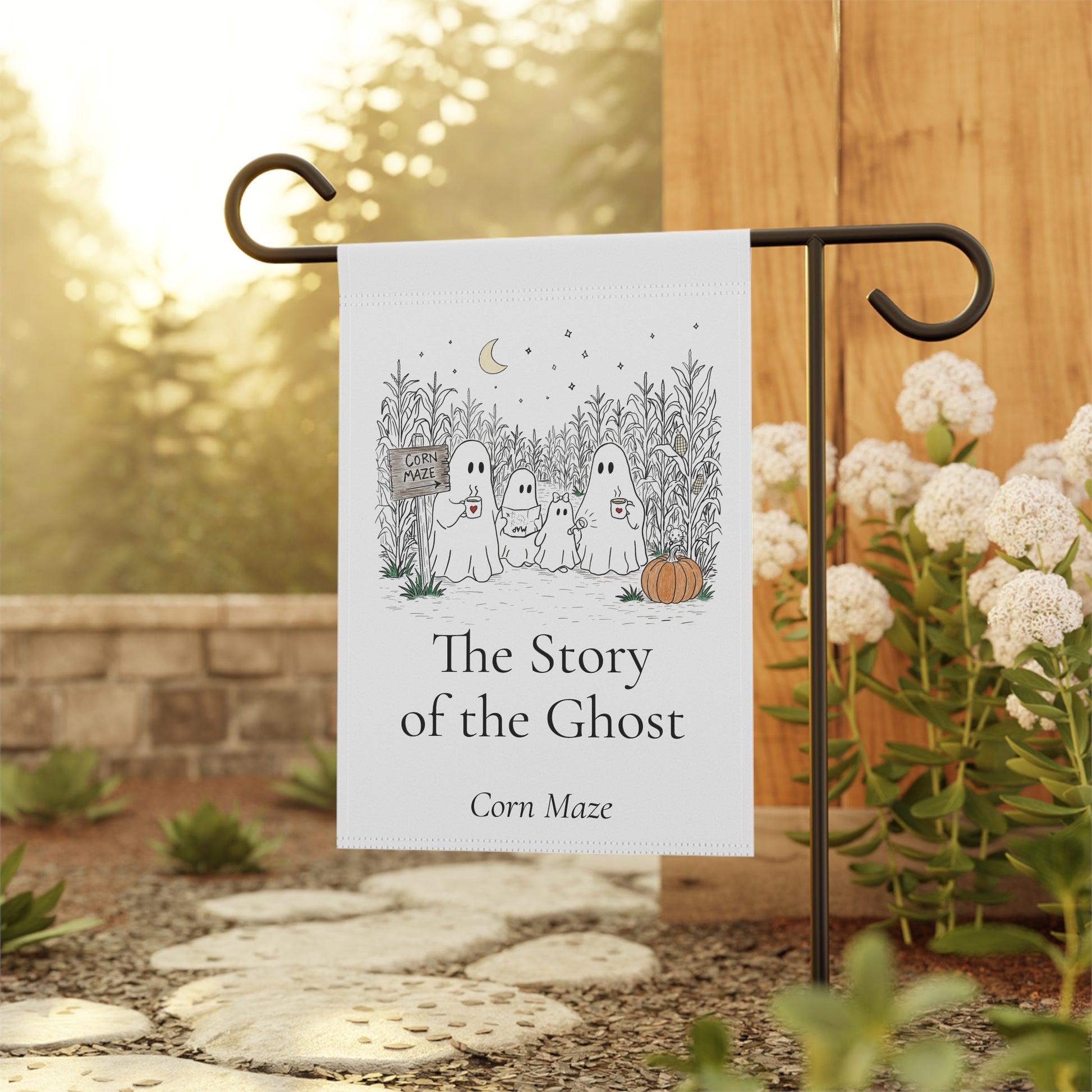 A vertical garden flag with a black metal stand, featuring a white background and a black line art illustration of three ghosts in a corn maze with the text 'The Story of the Ghost' and 'Corn Maze'. The flag is displayed outdoors in a garden setting with a wooden fence, stone path, and white flowers.