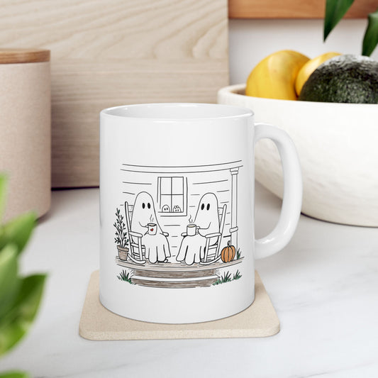 A white ceramic mug with a black line art illustration of two ghosts sitting on a porch, placed on a coaster in a kitchen setting.