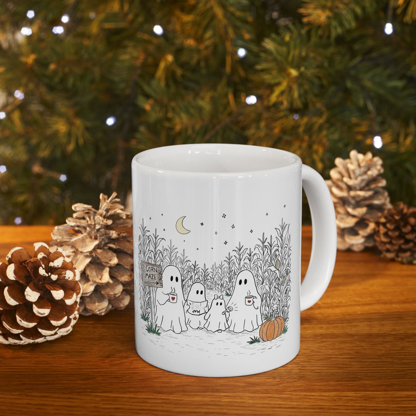 A white ceramic mug with a black and white cartoon ghost family illustration, set on a wooden table with pinecones and a blurred Christmas tree background.