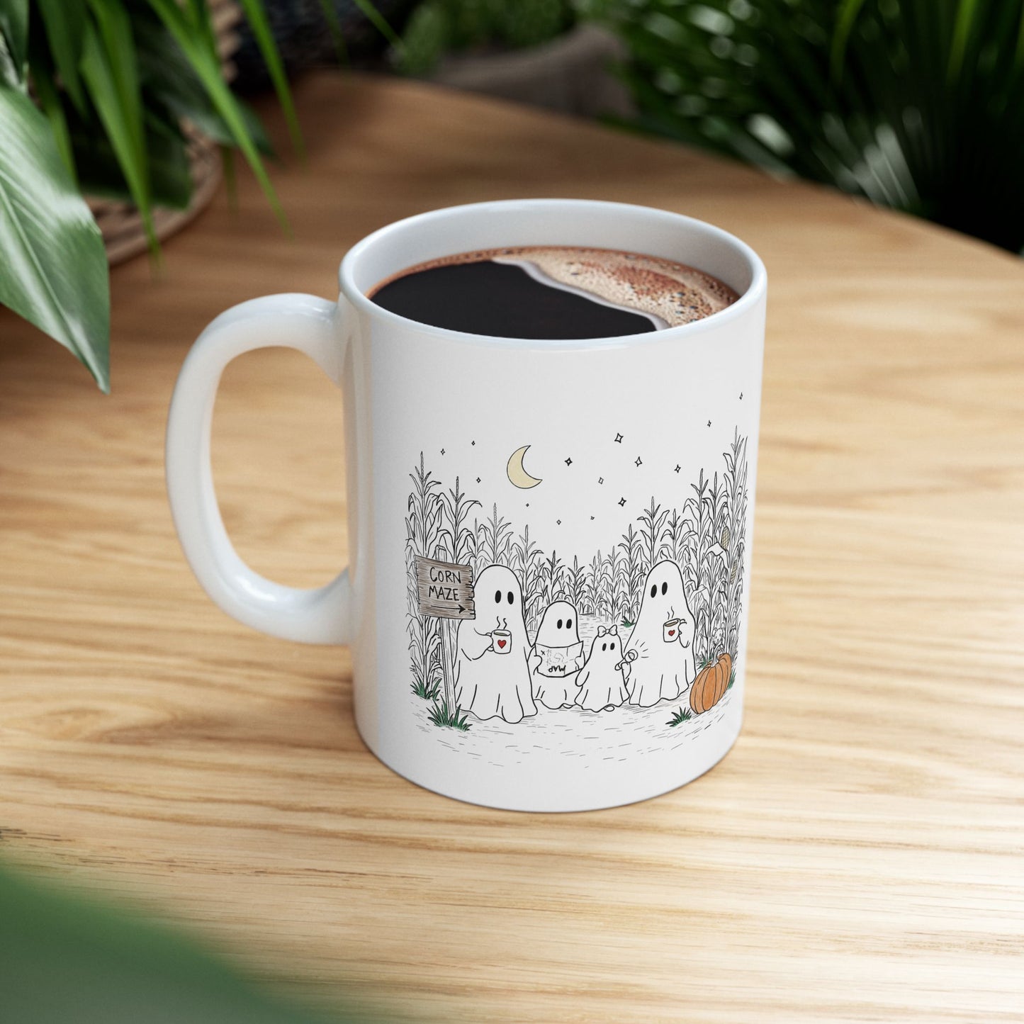 A white ceramic coffee mug with a cute cartoon ghost family illustration, filled with black coffee and placed on a light wooden table with blurred green plants in the background.