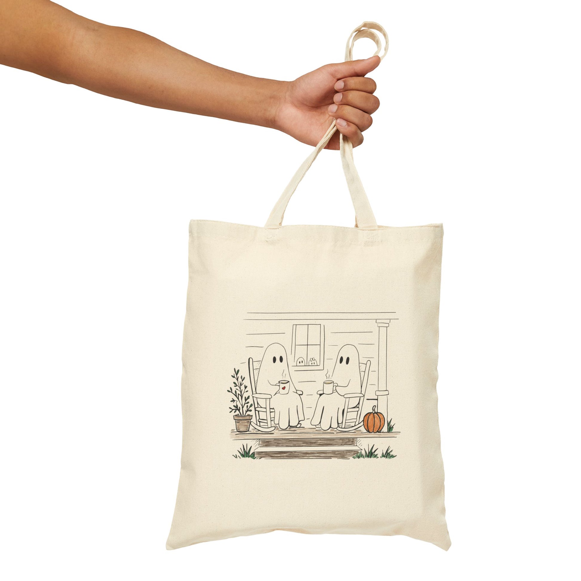 Natural canvas tote bag featuring a hand-drawn ghost family relaxing with coffee in rocking chairs on a front porch, with two little ghosts peeking through the window, a pumpkin, and subtle fall accents.