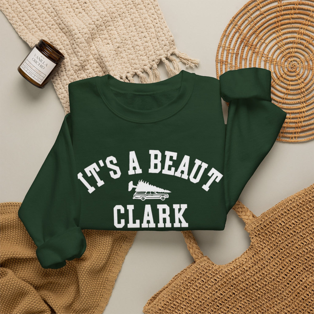 A dark green crewneck sweatshirt with white varsity-style text reading 'IT'S A BEAUT CLARK' and a small tree-truck graphic, styled in a flat lay with a cream knit blanket, woven placemat, tan knit fabric, and a small candle.