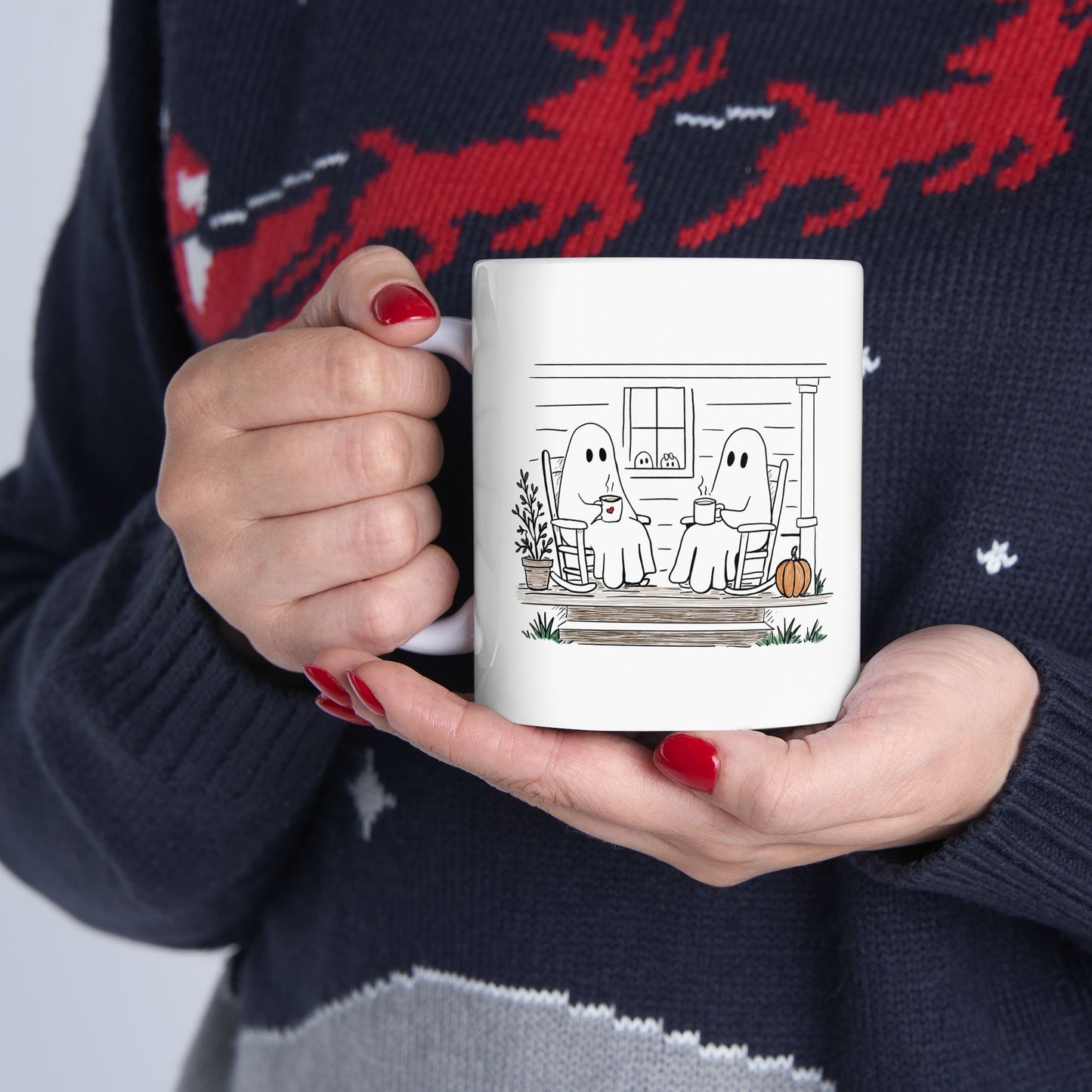 White ceramic mug featuring a hand-drawn ghost family sitting on a front porch in rocking chairs with coffee, with two little ghosts peeking through the window and subtle fall color accents.