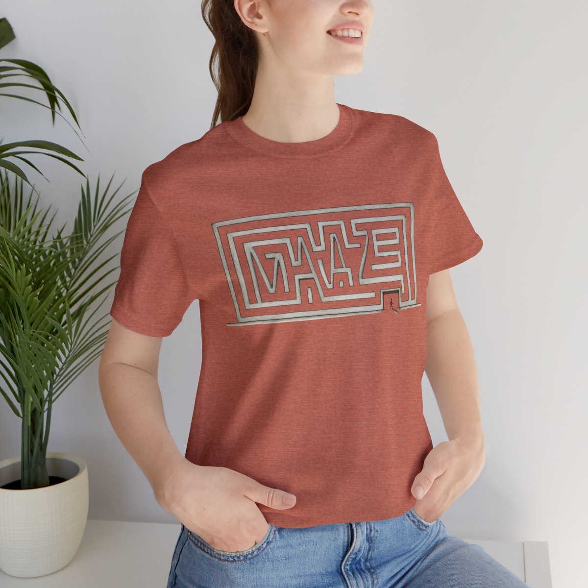 A woman wearing a heathered terracotta short-sleeve t-shirt with a white and gray geometric maze graphic on the chest, paired with blue jeans, seated next to a potted plant.