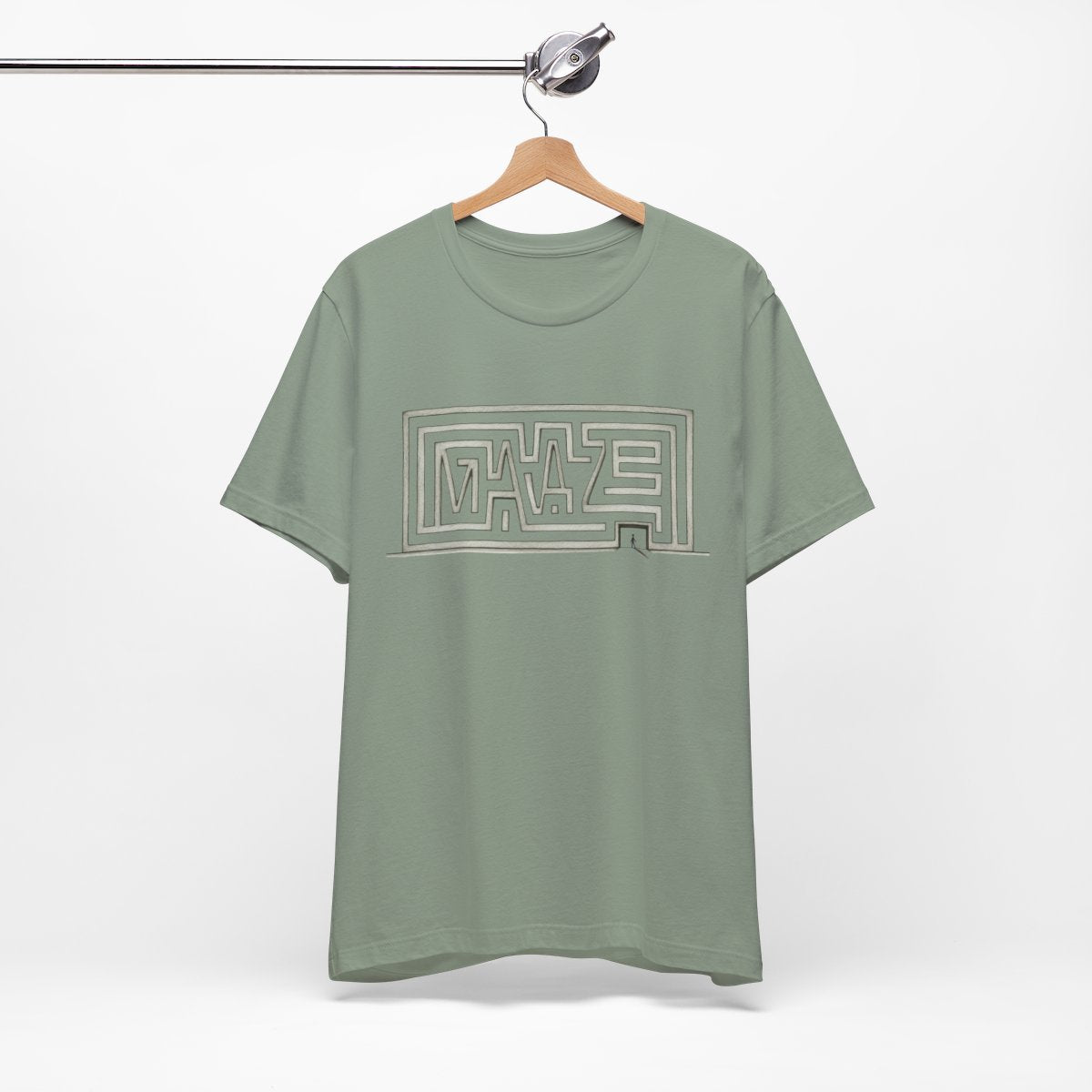 A sage green short-sleeve t-shirt hanging on a wooden hanger from a metal clothing rack, featuring a large geometric 'Maze' graphic printed on the chest.