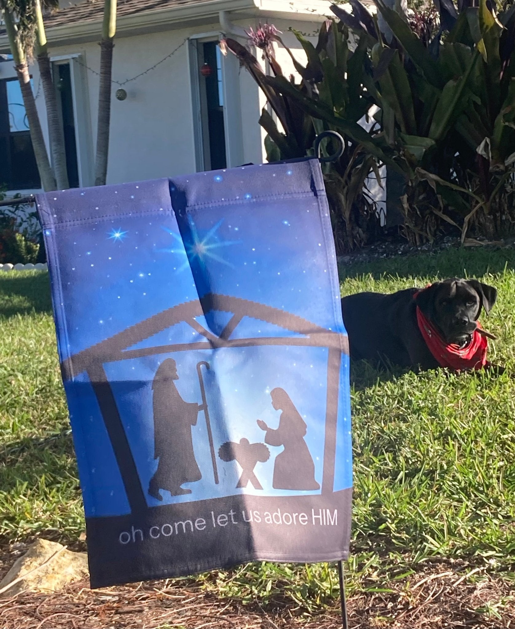 Decorative garden flag with nativity scene design on grass, with a dog in the background.