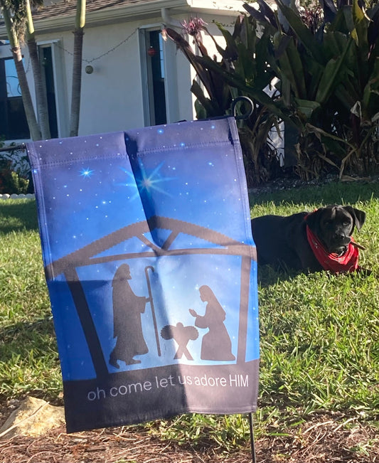 Decorative garden flag with nativity scene design on grass, with a dog in the background.