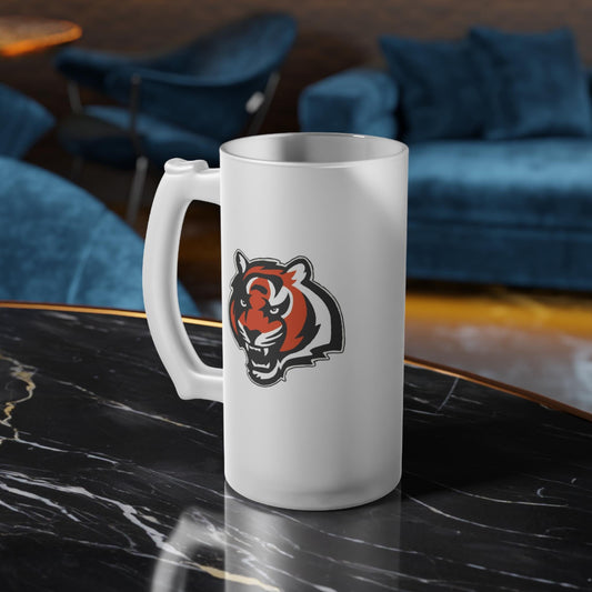 A white frosted glass beer mug with a hand-drawn tiger logo design, sitting on a marble surface with a blue chair and couch in the background.